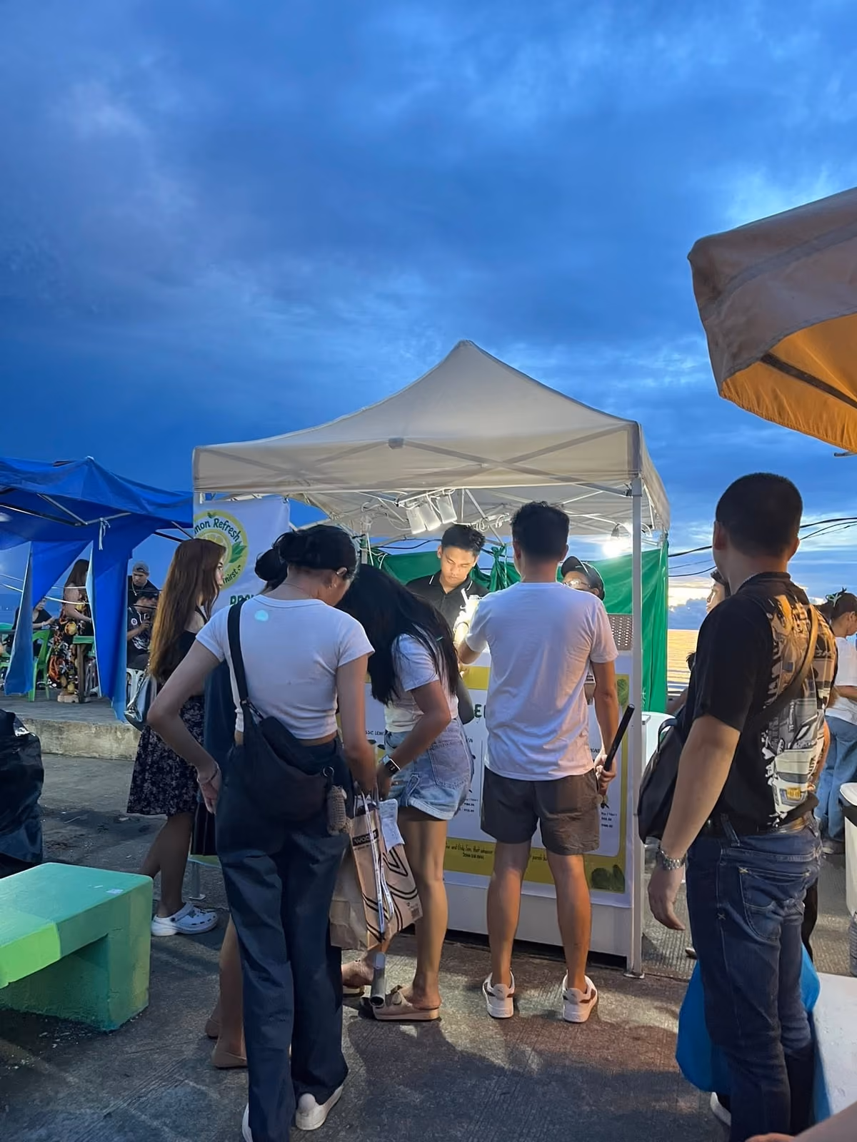 Customers gathered at the Lemon Refresh tent at an outdoor evening market, with the Lemon Refresh banner above the counter